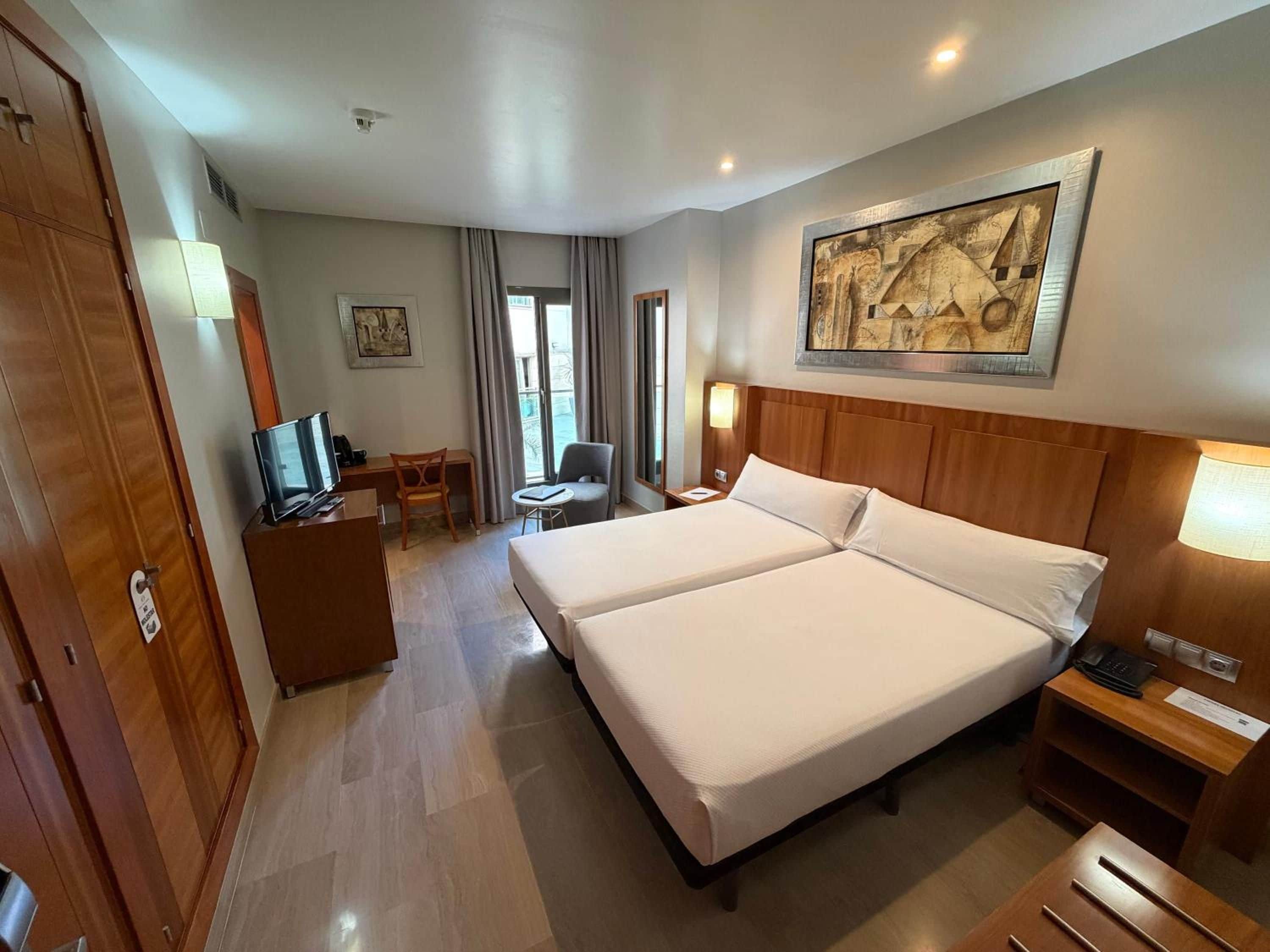 A hotel room with a double bed, a desk with a chair, a television, and a painting on the wall.