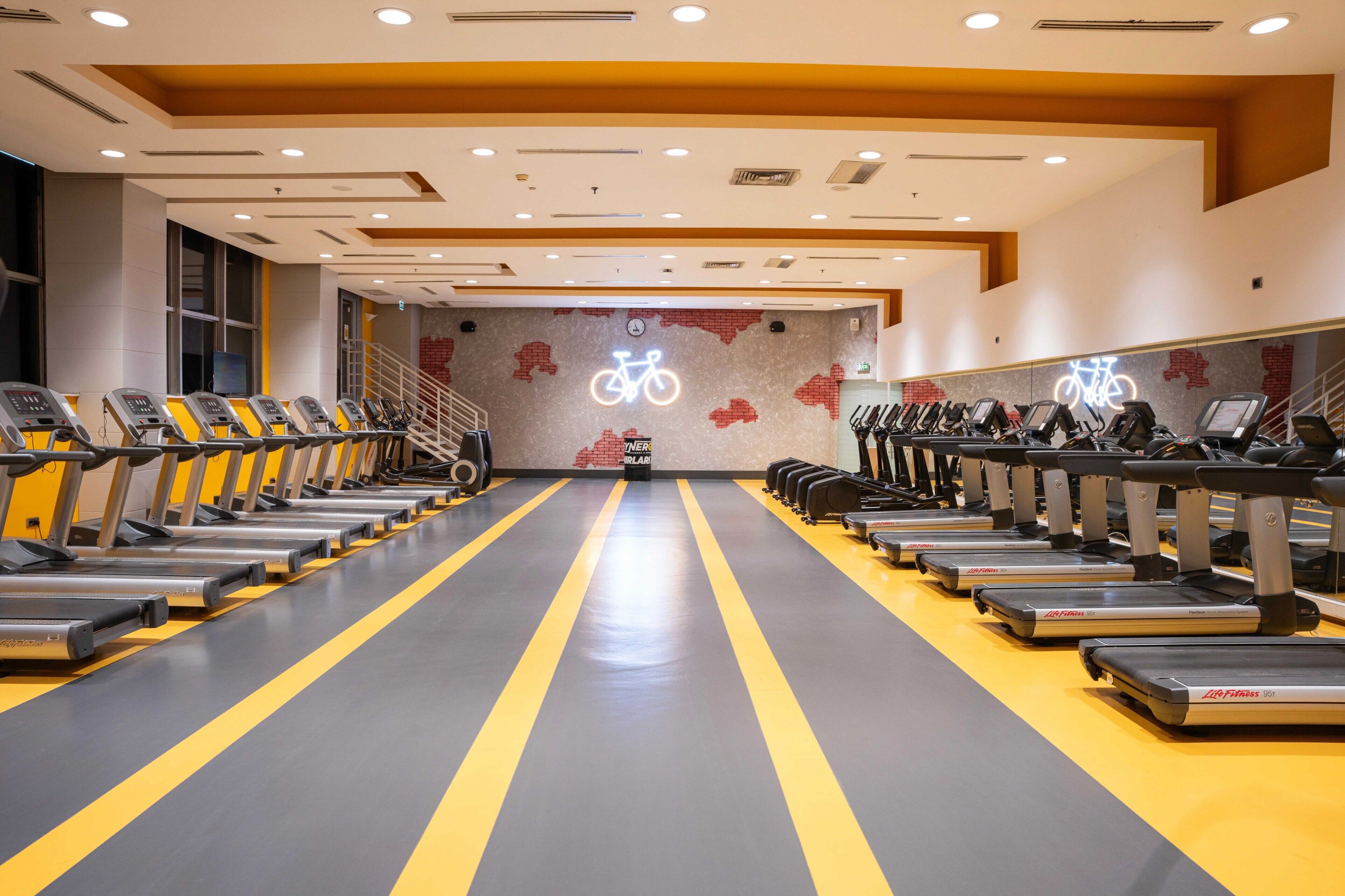 Ein Fitnessraum mit in Reihen angeordneten Laufbändern, einer Wand mit einem Fahrradmotiv und einer Treppe.