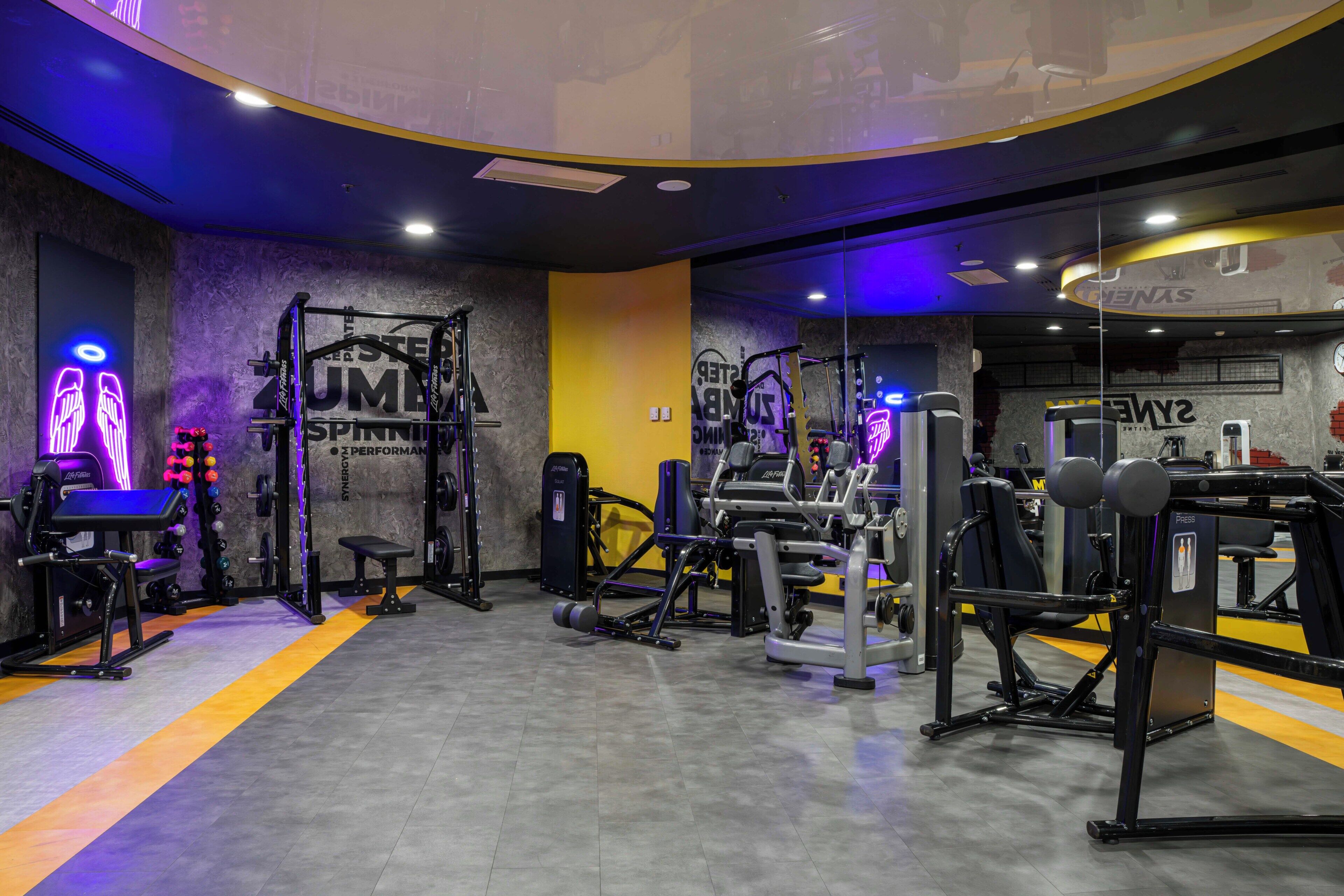 Une salle de sport équipée de divers appareils d’exercice, d’une horloge murale et de motifs inspirants sur les murs.