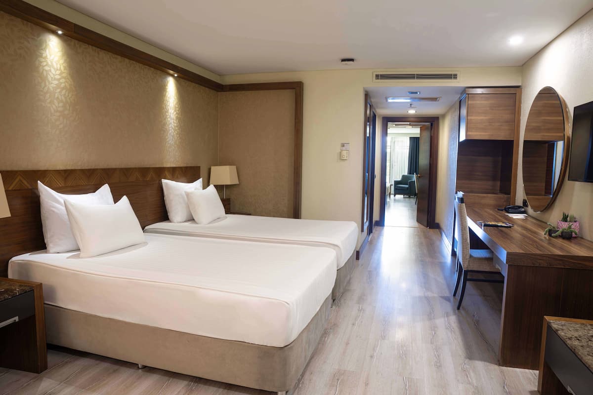 A hotel room with two beds, a desk, a chair, a mirror, and a hallway leading to another room.