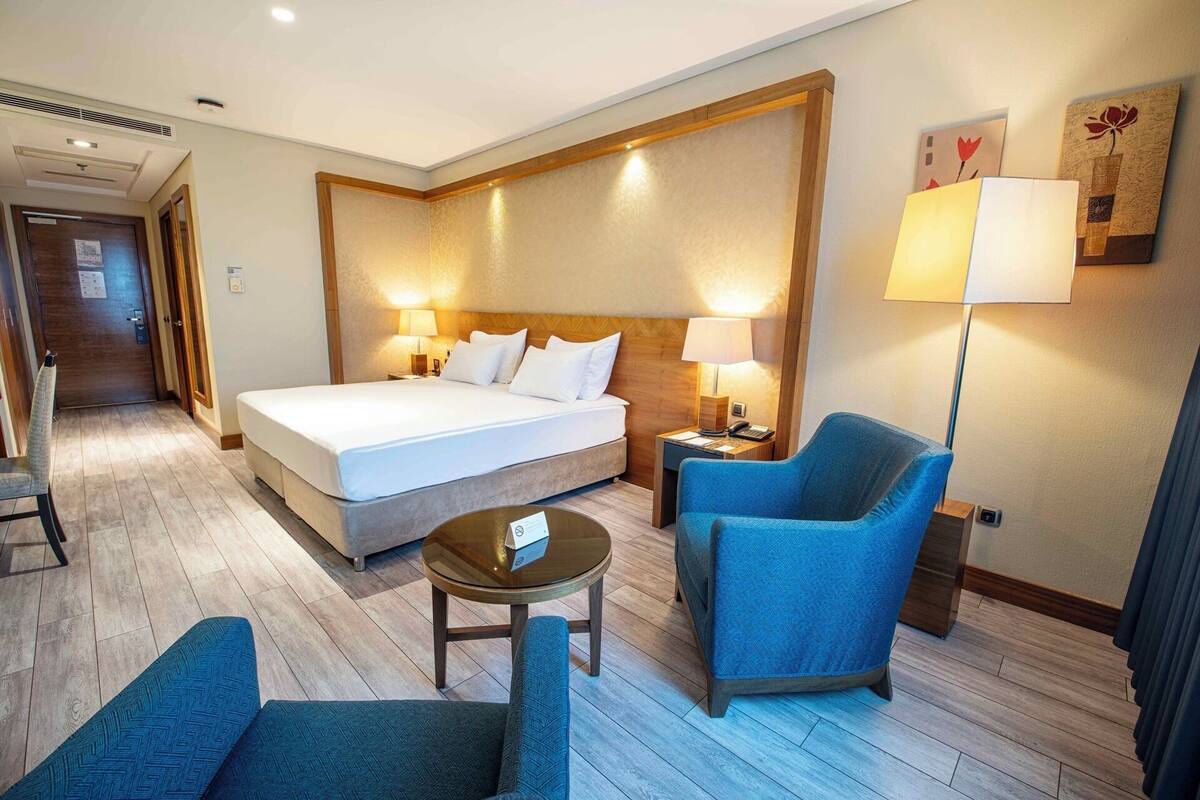 A hotel room with a bed, two bedside lamps, a chair, a small table, and a door.