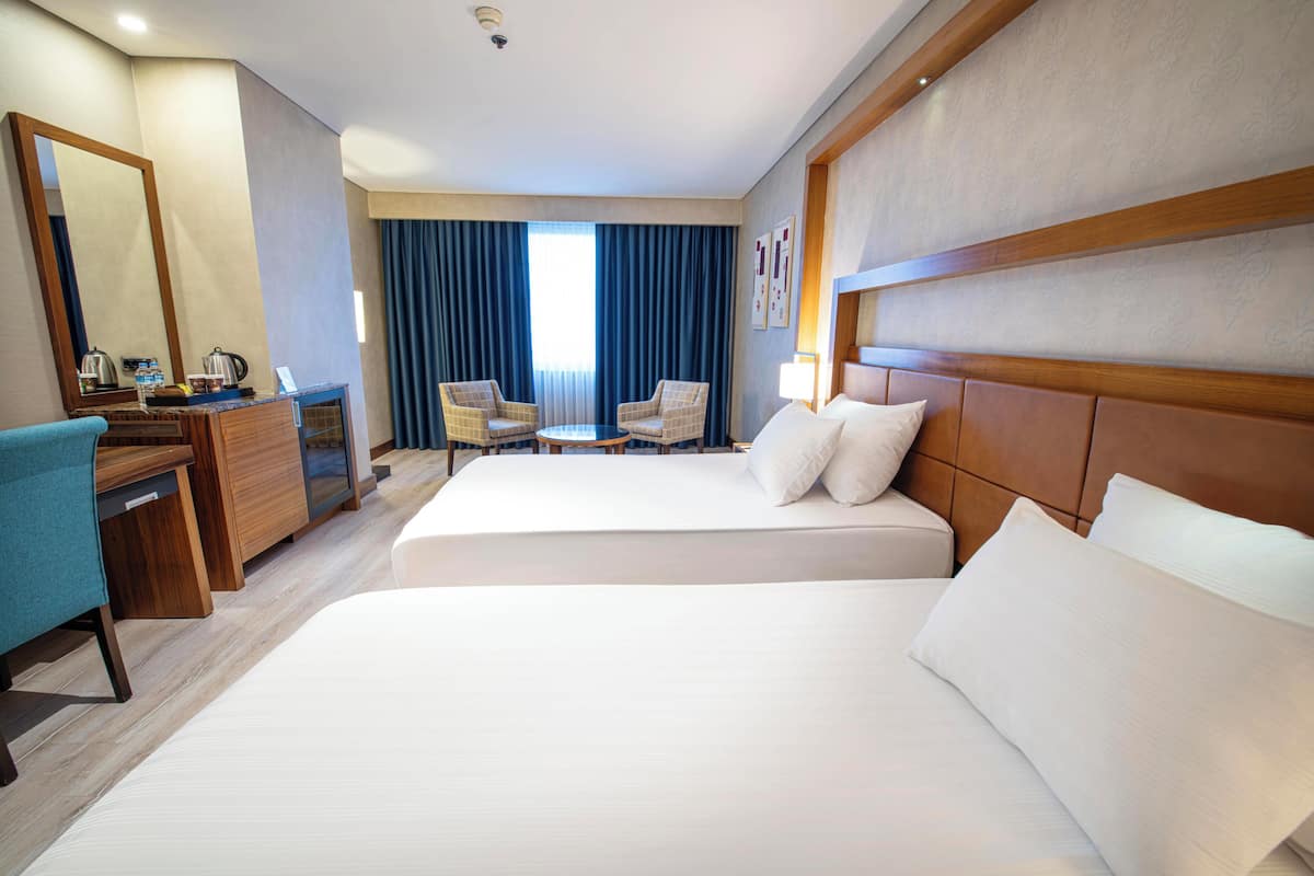 A hotel room with two beds, a desk with a chair, a small table, and a large mirror.