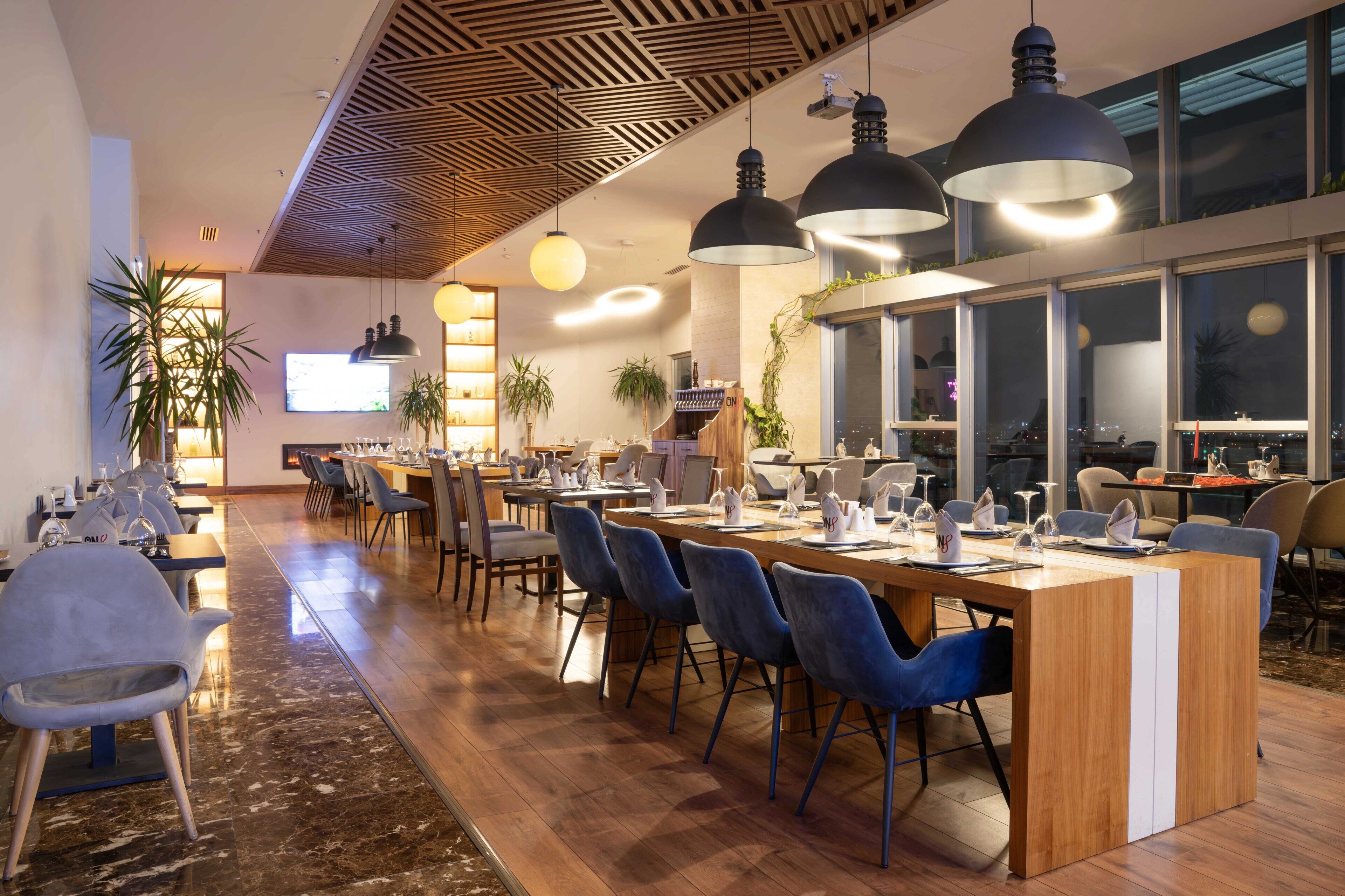 Un restaurante moderno con una larga mesa común, sillas azules, iluminación colgante y ventanas amplias.