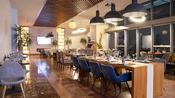 Un restaurante moderno con una larga mesa común, sillas azules, iluminación colgante y ventanas amplias.