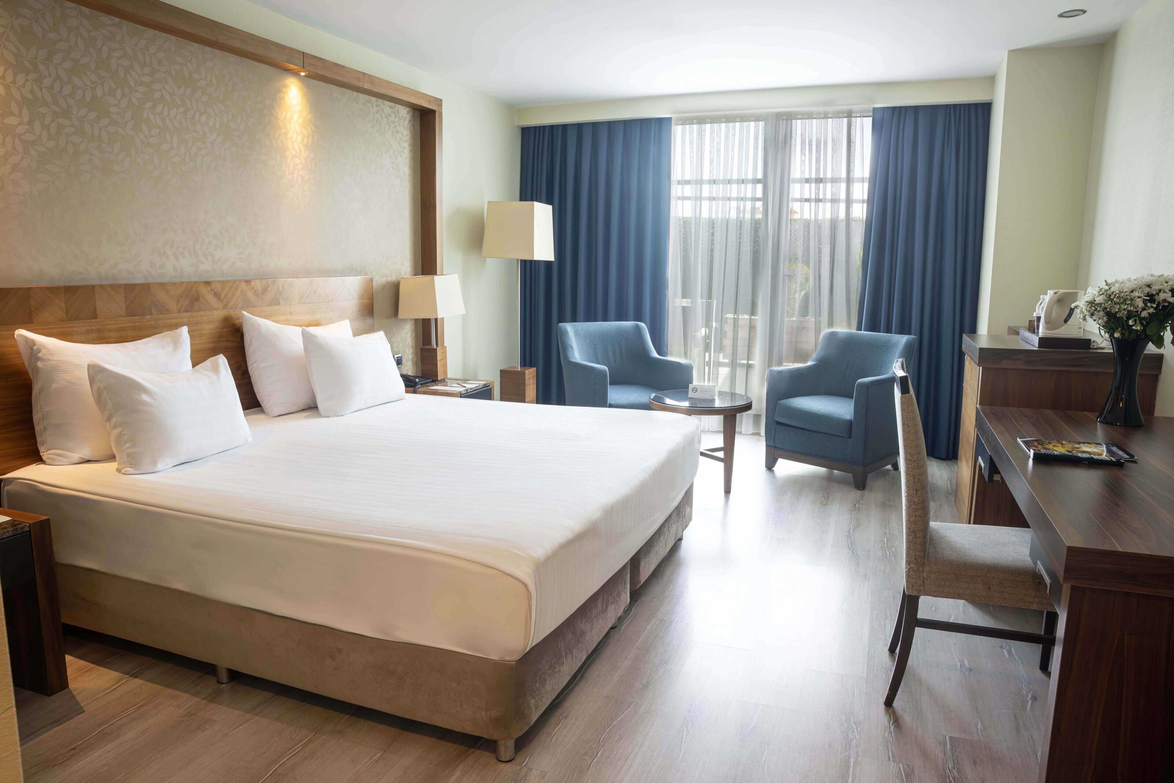 A hotel room with a large bed, two armchairs, a desk, a lamp, and a view of the outdoors.