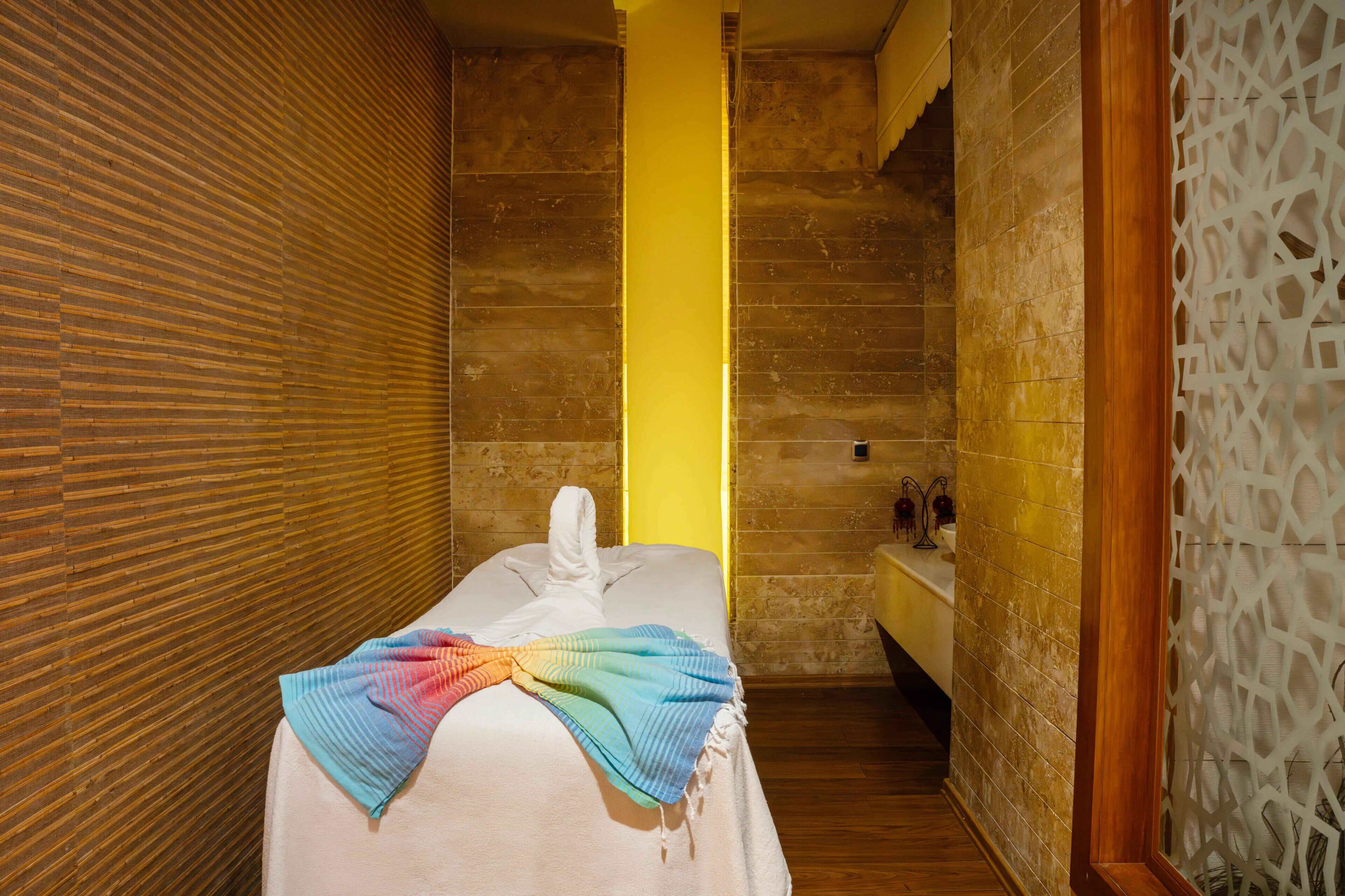 A spa room with a wooden slat wall, a yellow pillar, a bed with a colorful towel, and a white partition.