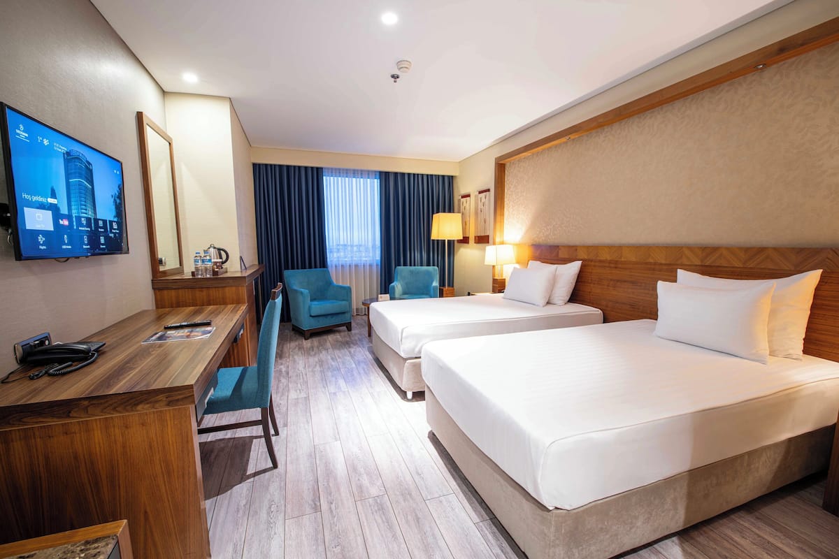 A hotel room with two beds, a desk with a computer, a TV mounted on the wall, and a large window with curtains.