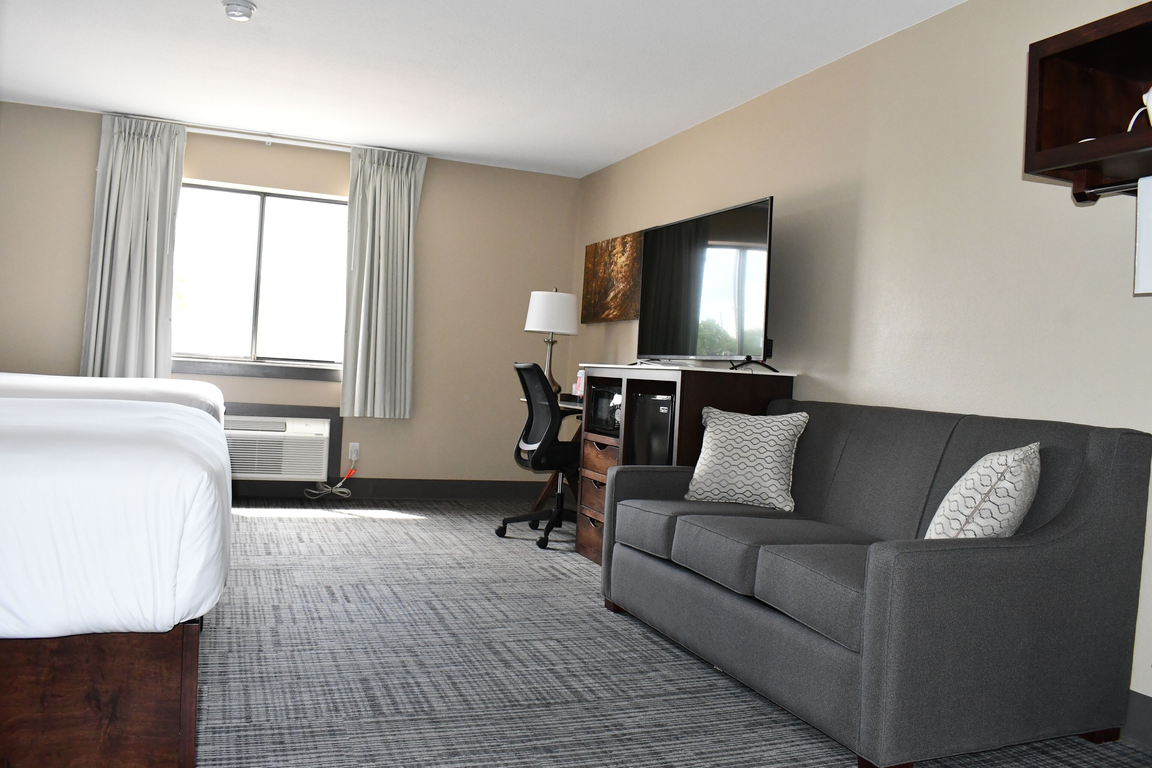 A hotel room with a bed, a grey sofa, a desk with a chair, a flat-screen TV, and a window with curtains.