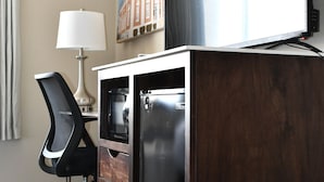 A hotel room with a wooden desk, a black office chair, a lamp, a refrigerator, and a television on a stand. - Cobblestone Inn & Suites - Trenton (Trenton)