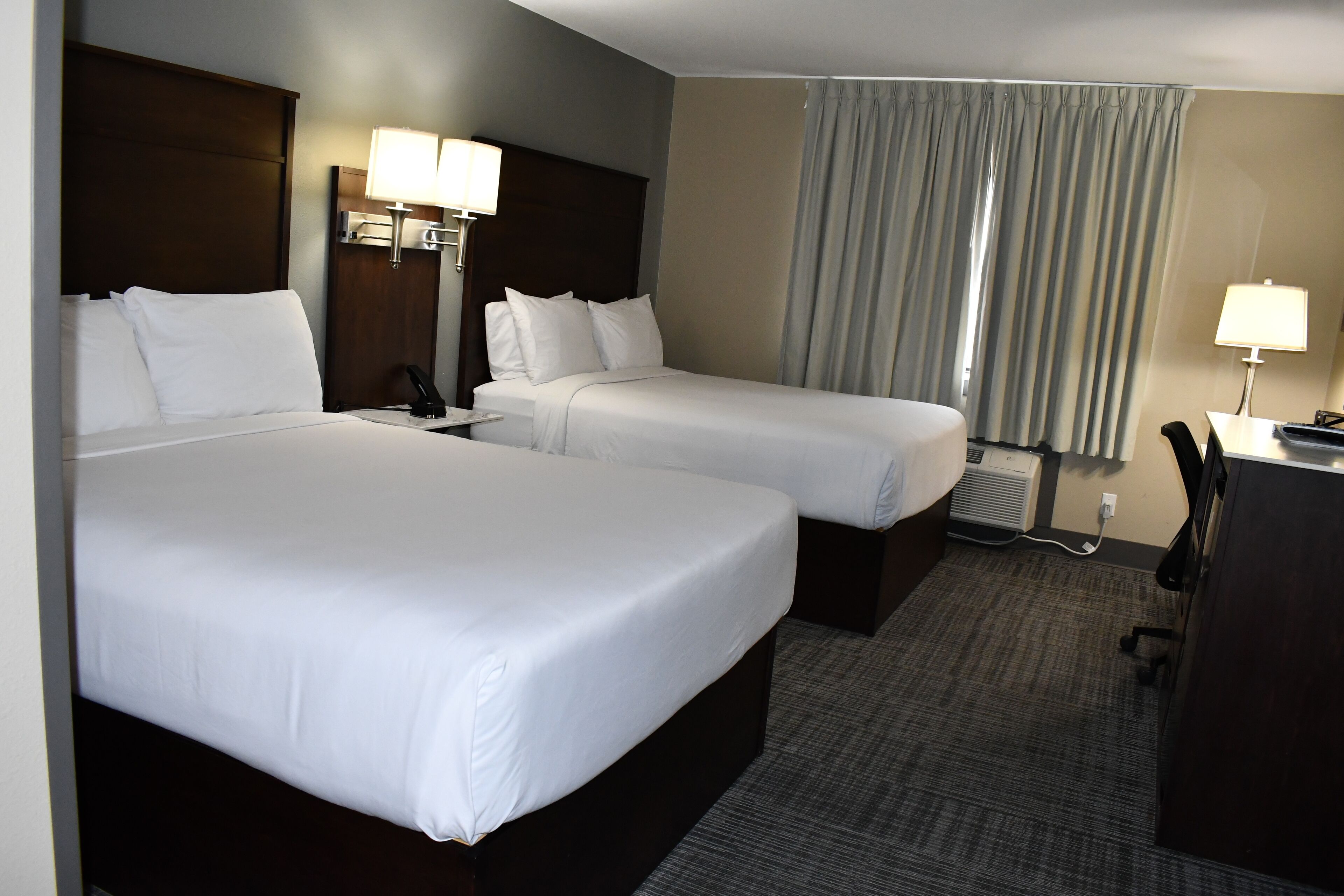 A hotel room with two beds, a desk, a chair, a lamp, and a window with curtains.
