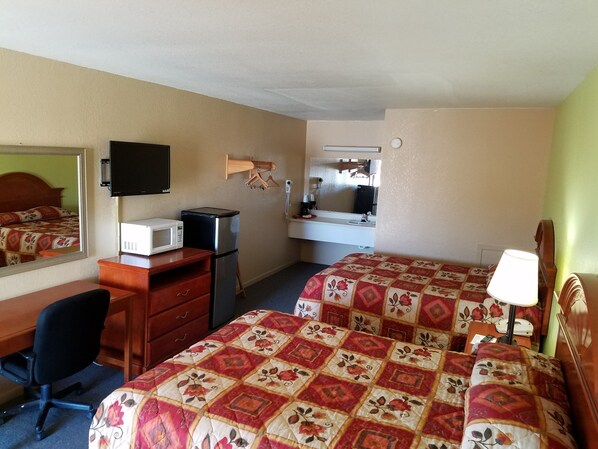 A hotel room with two beds, a desk with a computer, a refrigerator, a microwave, a TV, a mirror, and a bedside table with a lamp.