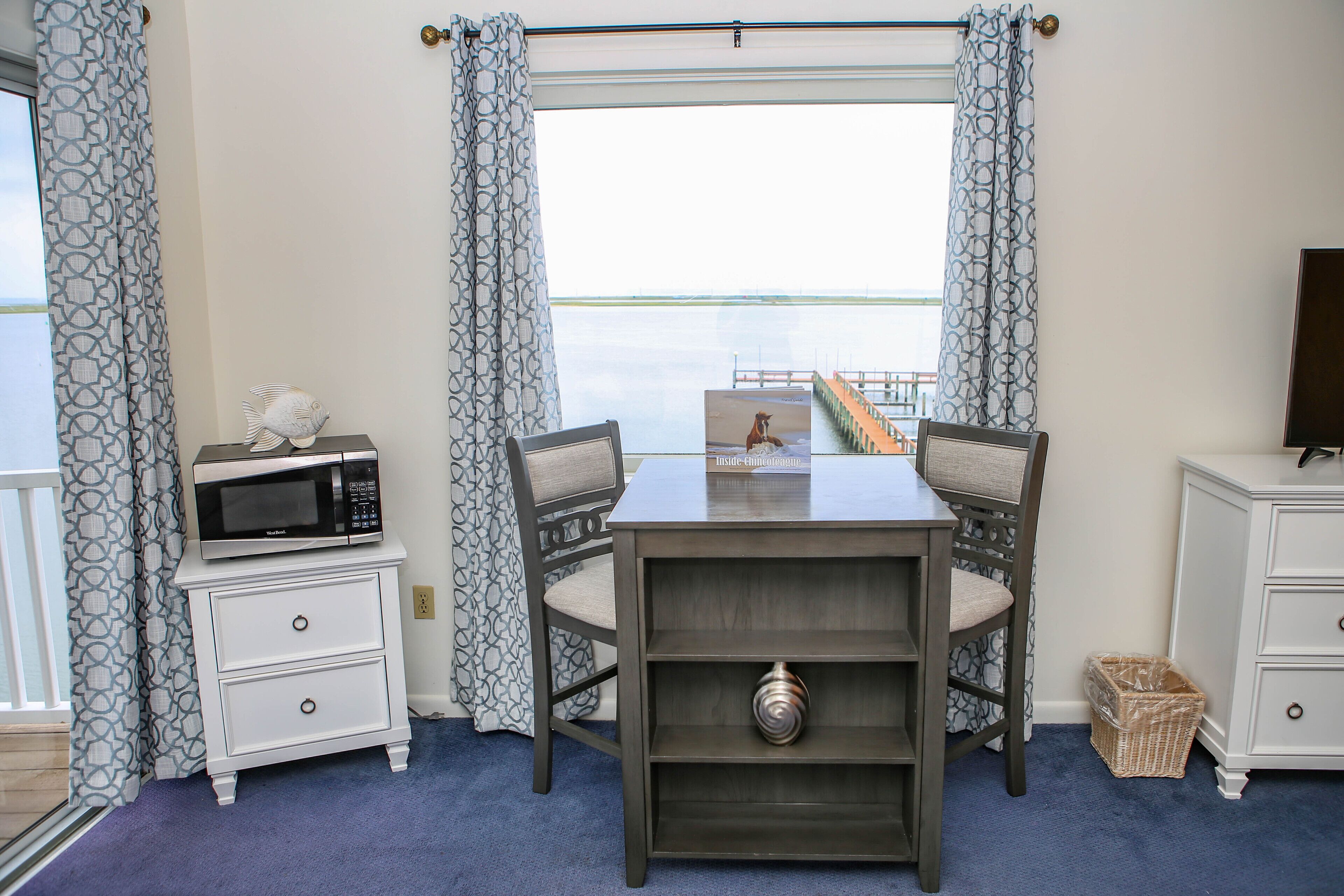 A room with a table and chairs, a microwave on a nightstand, a wicker basket, and a view of the water through the window.