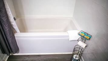A bathroom with a white bathtub, a dark wooden floor, a white towel, and a metal basket with a toilet paper roll.
