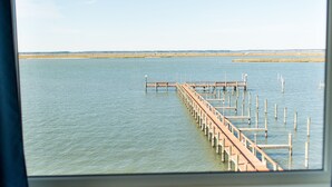 Vista de un muelle que se adentra en aguas tranquilas, tomada desde una habitación con una mesa y un florero.