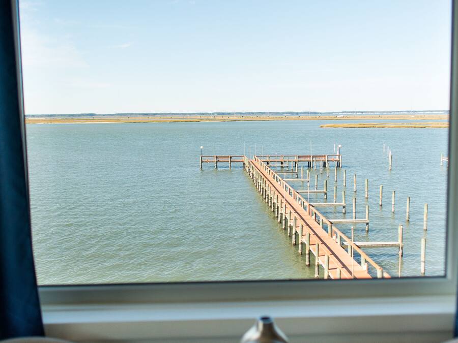 Vista de un muelle que se adentra en aguas tranquilas, tomada desde una habitación con una mesa y un florero.
