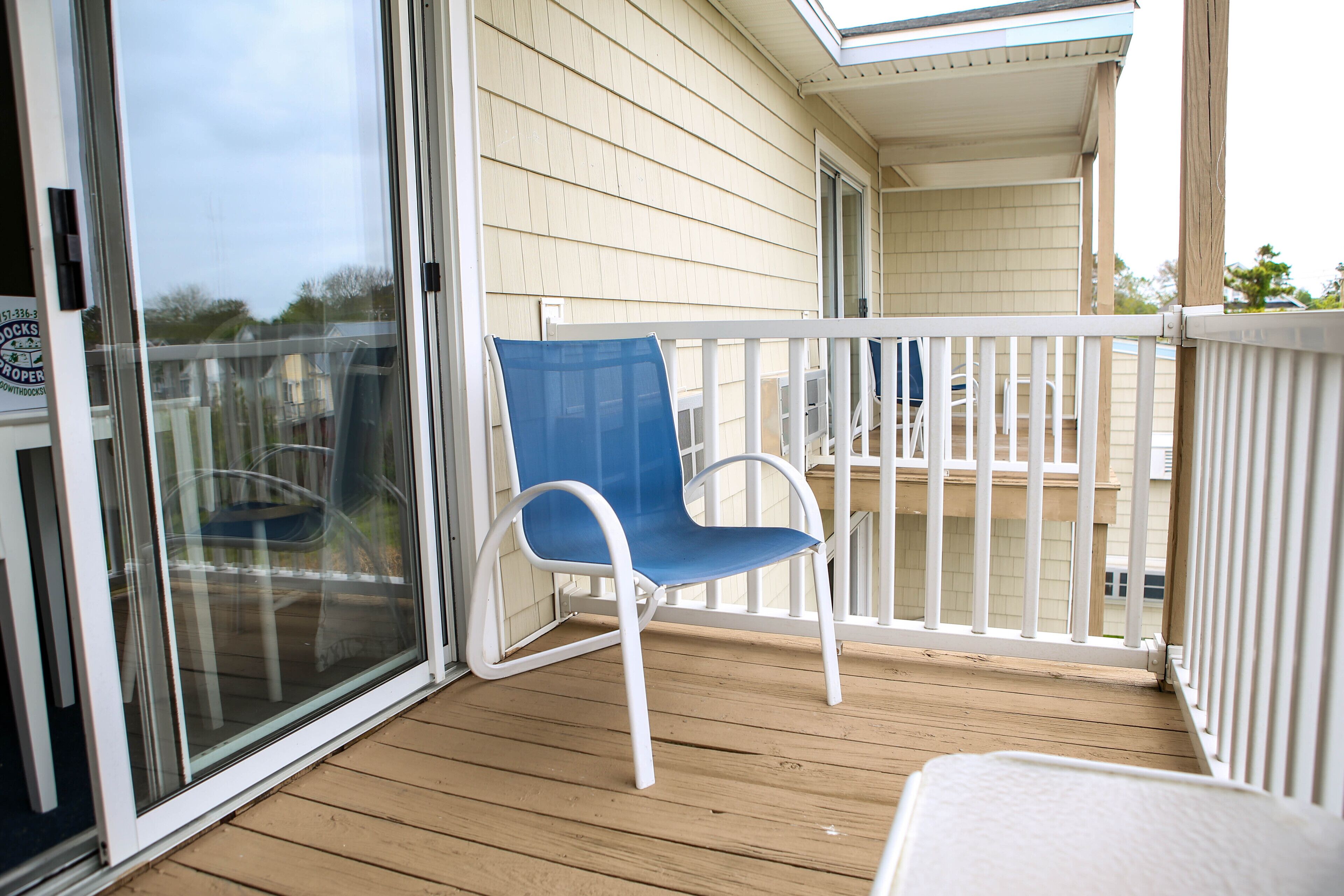 A wooden deck with a blue chair and a white table, a glass door, and a covered porch area.