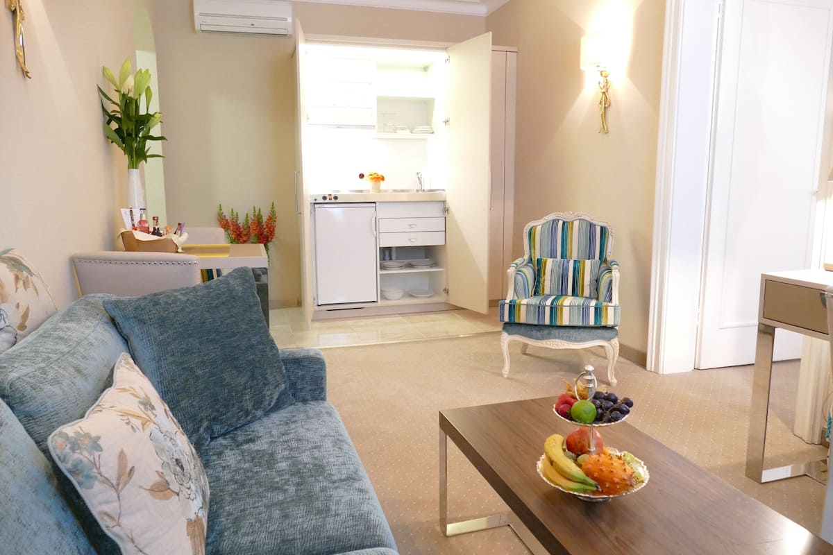 A living room with a sofa, a coffee table with fruit, a striped armchair, and a kitchen in the background.