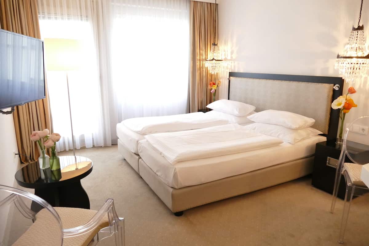 A hotel room with a double bed, a TV, a desk with a chair, a small table with a vase of flowers, and a chandelier.