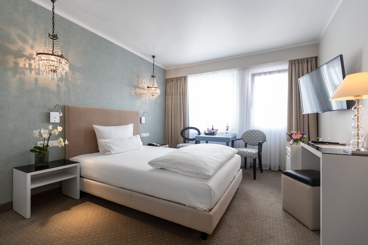 A hotel room with a large bed, bedside table, desk, chair, TV, and a chandelier.