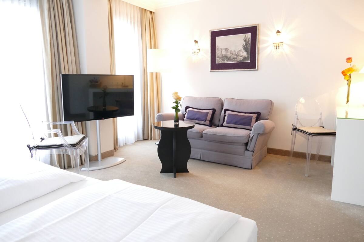 A hotel room with a bed, a grey sofa, a TV, a small table, and a chair.