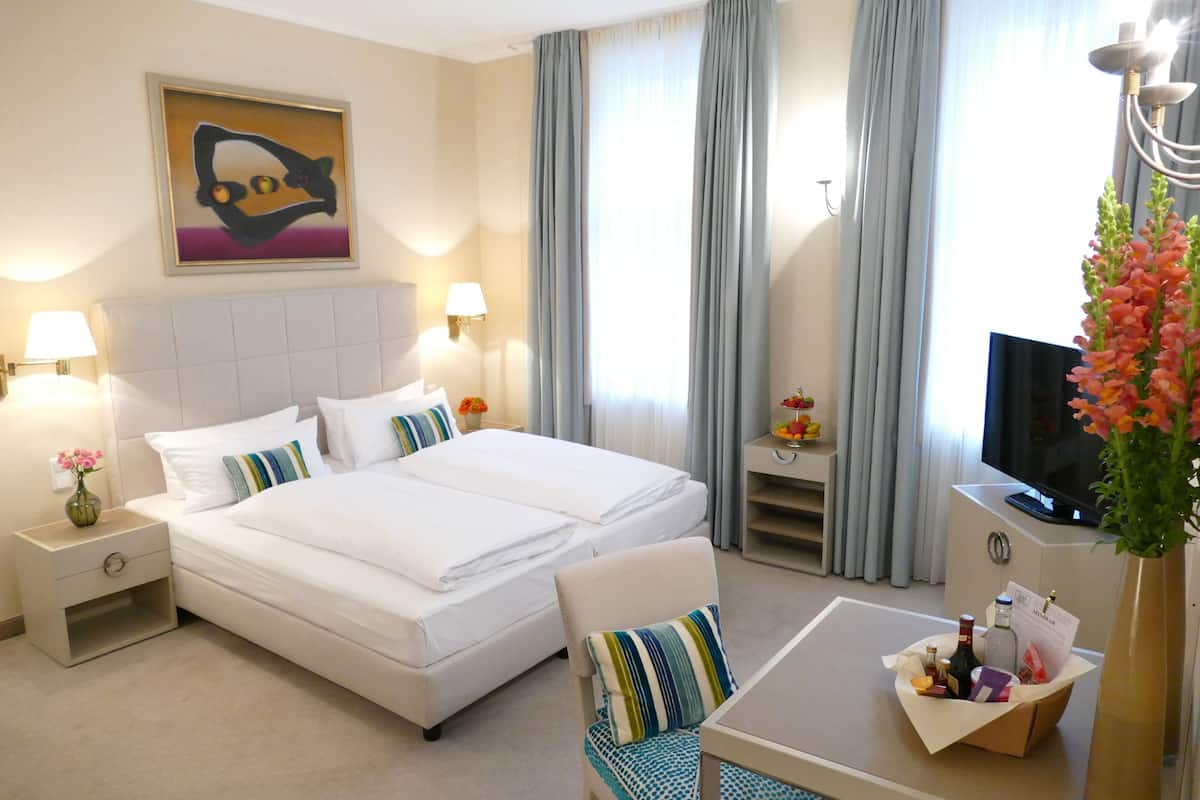 A hotel room with a large bed, a desk with a flat-screen TV, a vase with flowers, and a painting on the wall.