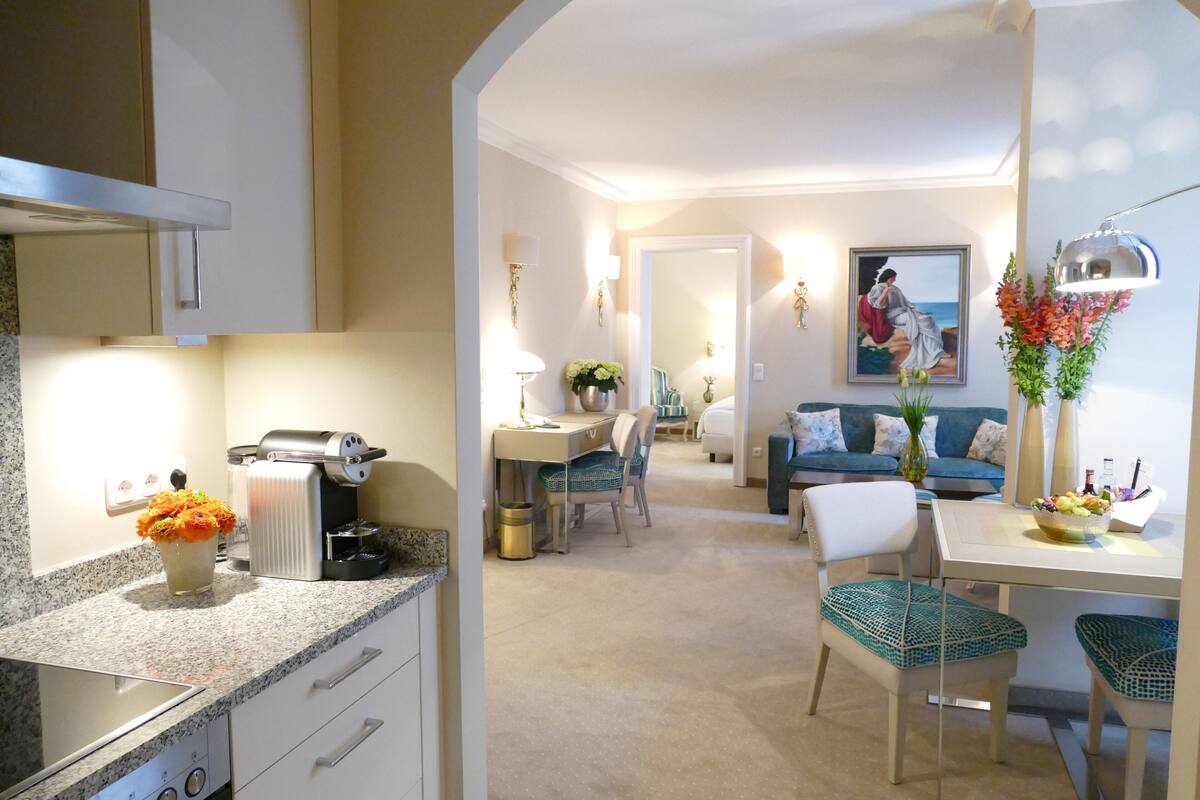 A modern kitchen with a coffee maker, a dining area with a table and chairs, and a living room with a sofa and a painting on the wall.
