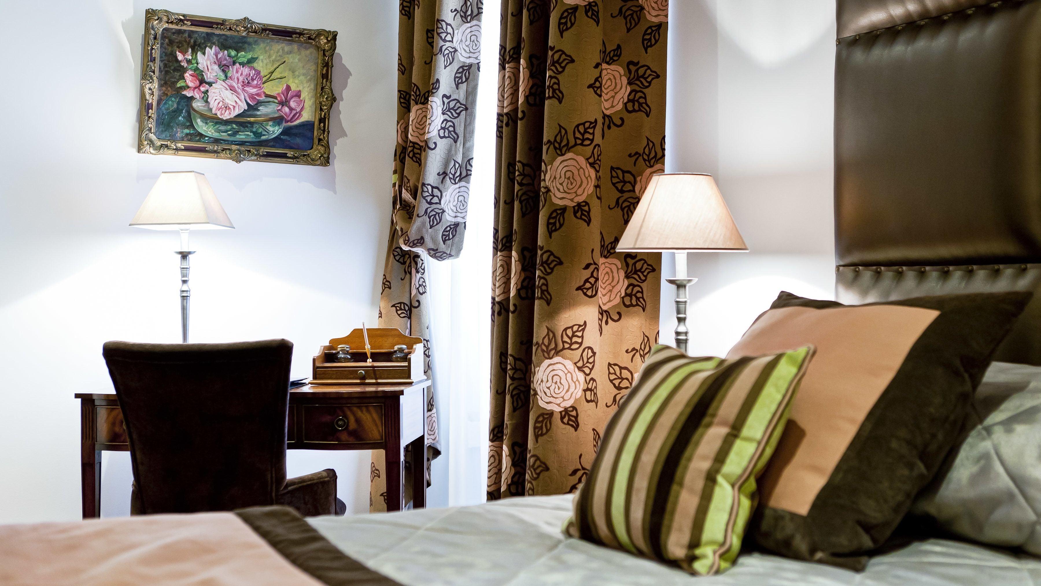 A hotel room with a bed, two bedside lamps, a desk with a telephone, a chair, and a painting on the wall.
