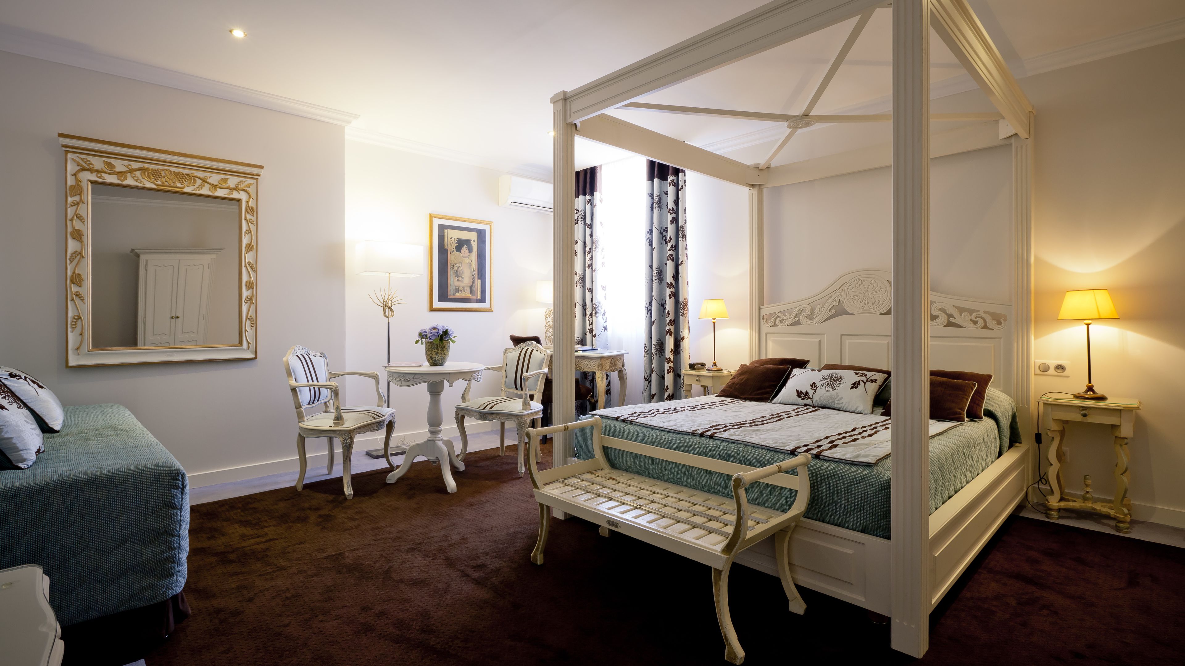 Una cama con dosel y cuatro columnas cubierta con una colcha verde y blanca, una pequeña mesa con sillas, un espejo con marco decorativo y una lámpara en una mesita de noche.