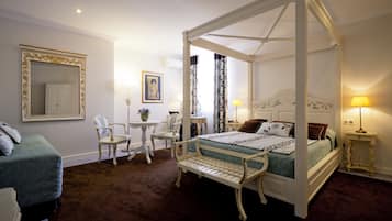 Una cama con dosel y cuatro columnas cubierta con una colcha verde y blanca, una pequeña mesa con sillas, un espejo con marco decorativo y una lámpara en una mesita de noche.