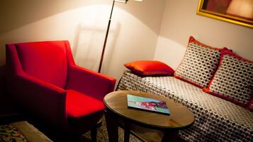 Une pièce avec un fauteuil rouge, un canapé à motifs, une petite table sur laquelle se trouve un magazine et un lampadaire.