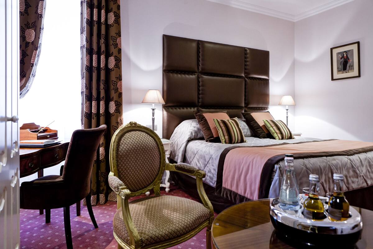 A hotel room with a large bed, a desk, a chair, and a bedside table with bottles.