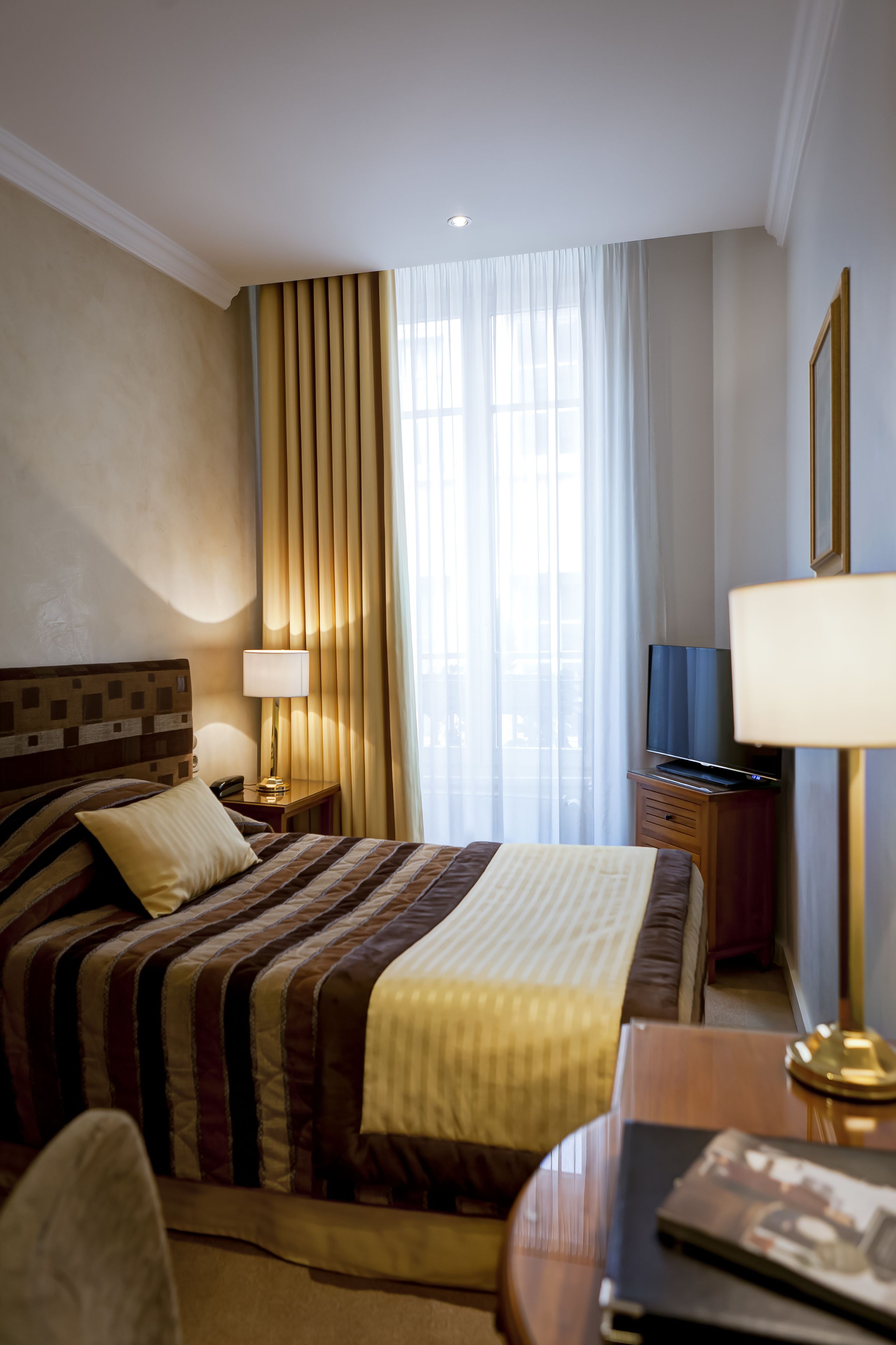 A hotel room with a bed, a bedside table with a lamp, a TV, and a window with curtains.