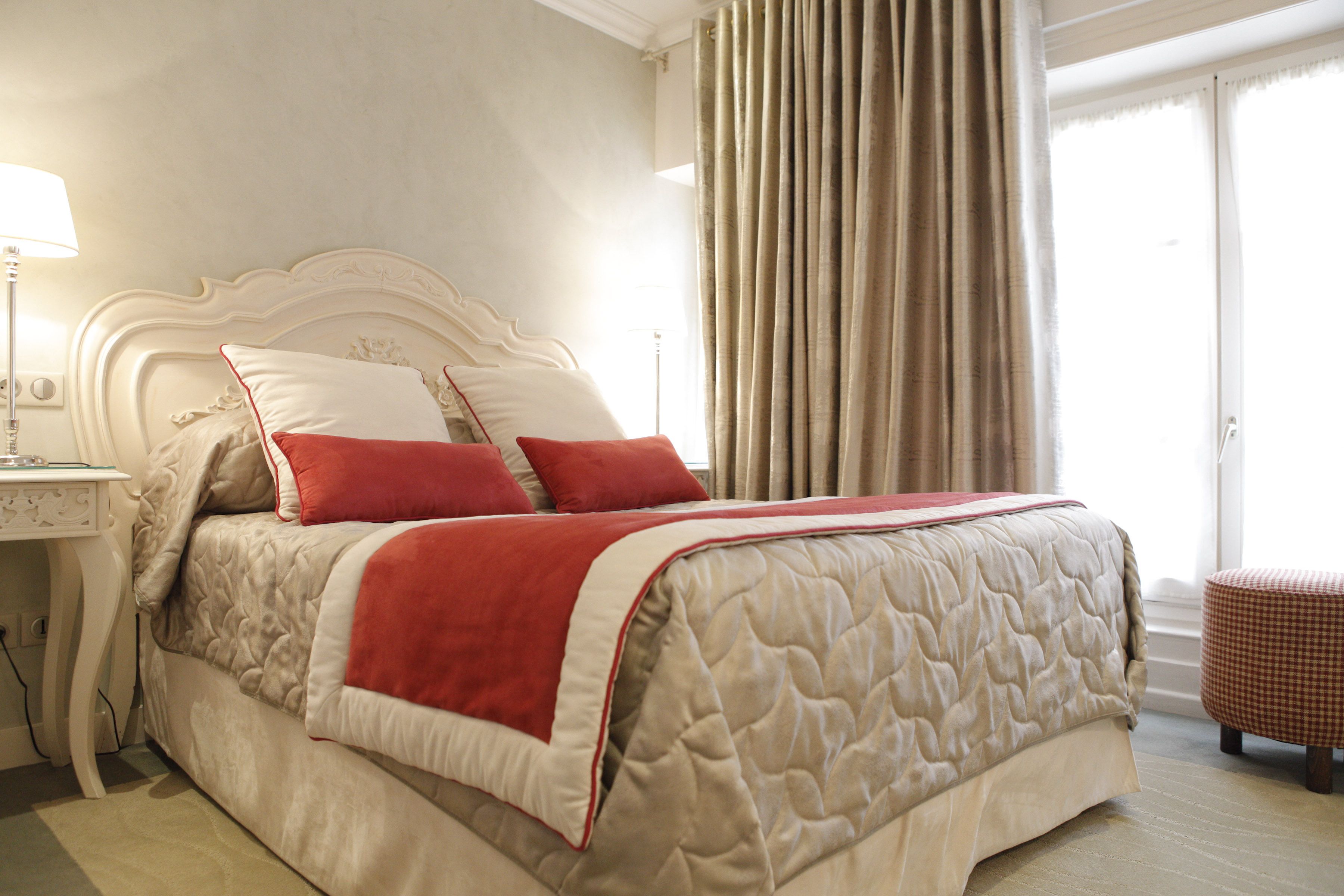Una cama bien hecha con cubrecolcha acolchado, una mesita de noche con lámpara y una ventana con cortinas.