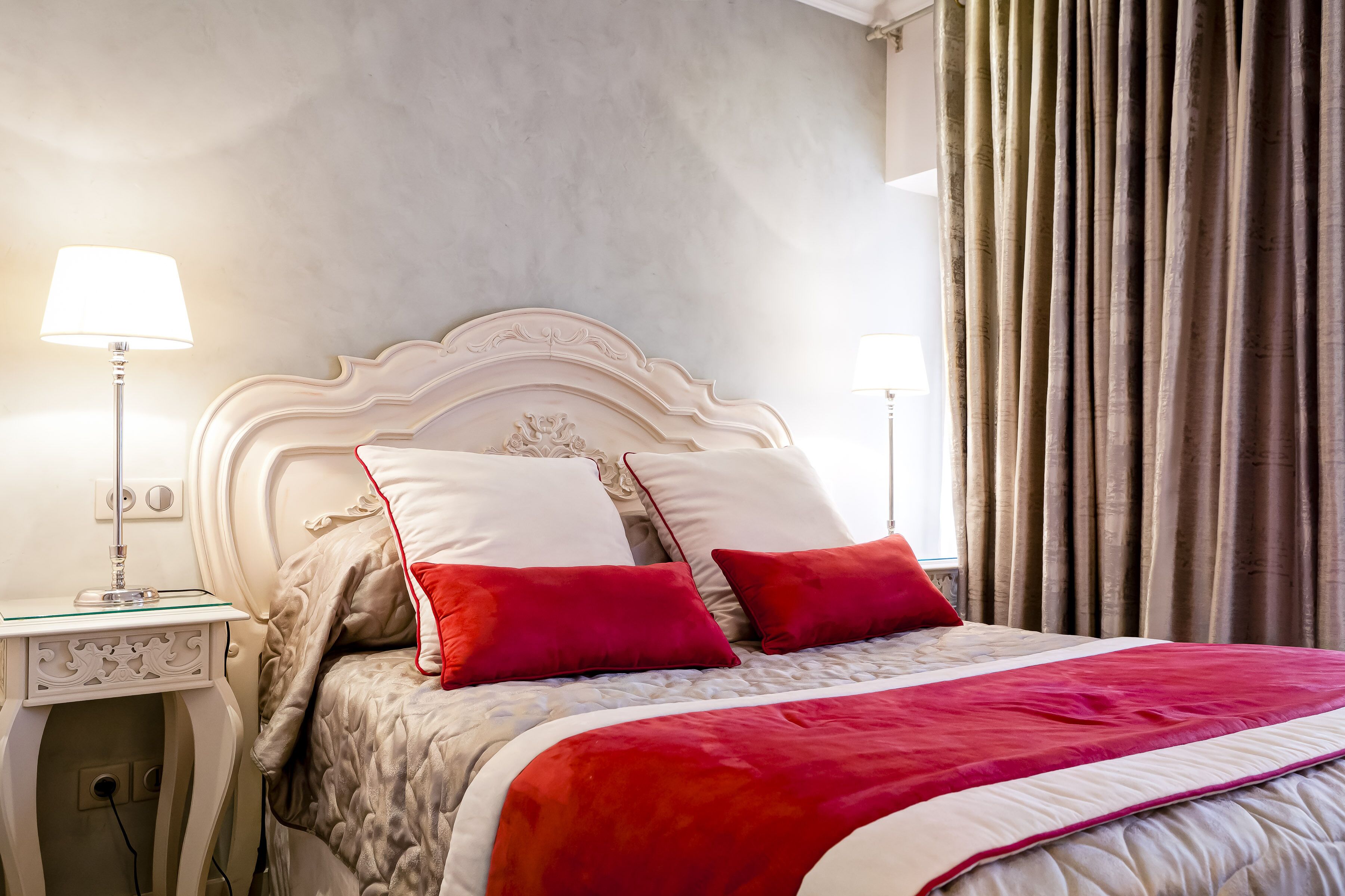A neatly made bed with a white headboard, red and white pillows, and a red and white bedspread. There are two bedside tables with lamps and a small decorative item on one.