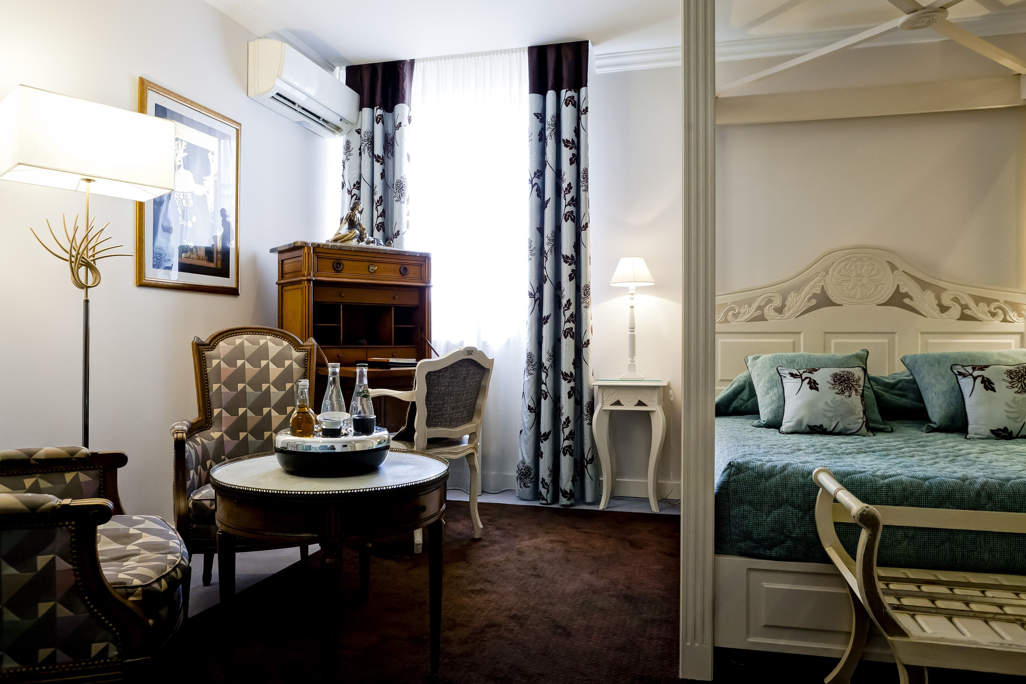 A hotel room with a bed, a desk with a lamp, a chair, a small table with a bottle, and a picture on the wall.