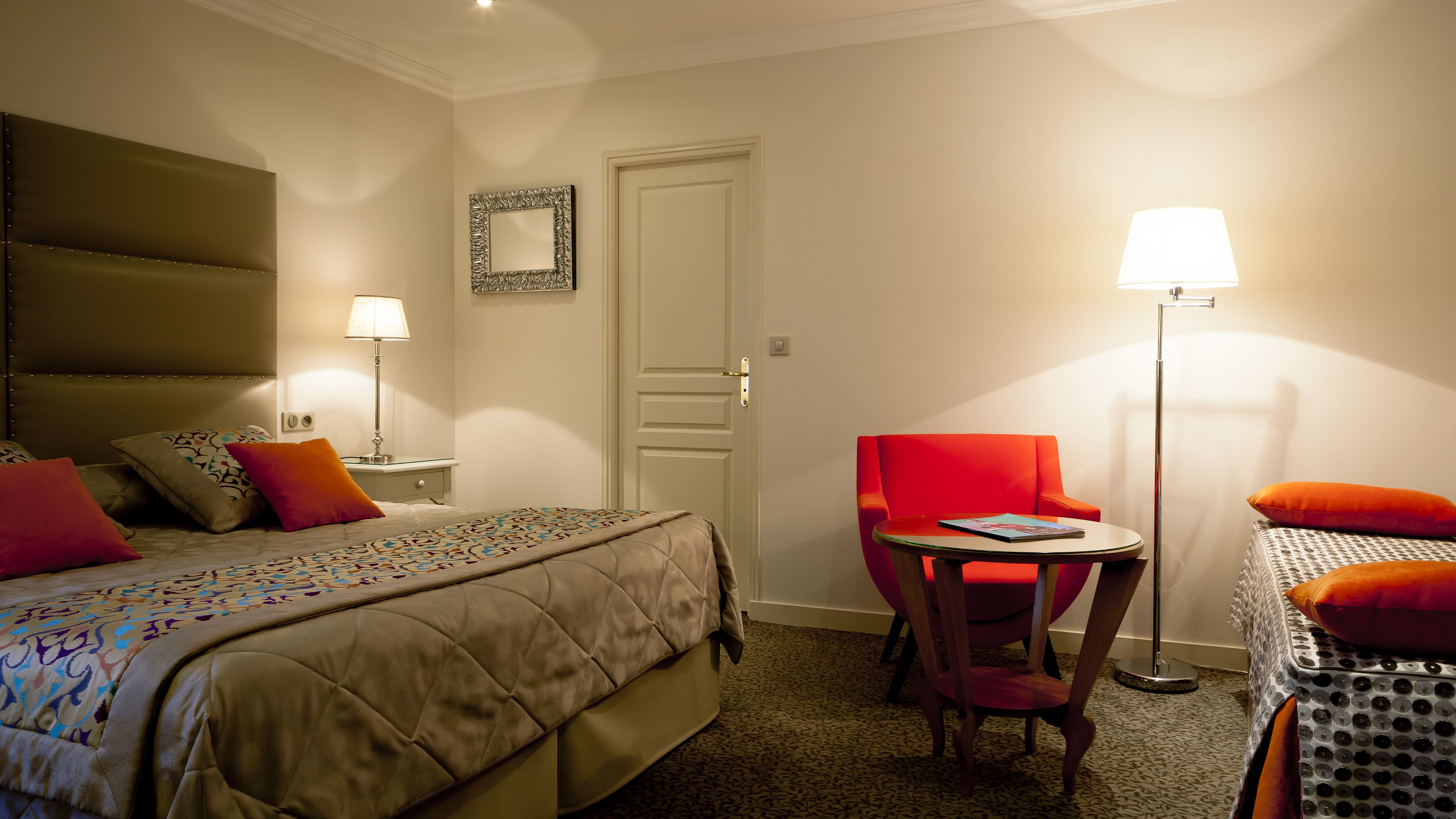 A hotel room with a bed, a bedside table with a lamp, a chair, and a small table with a magazine.