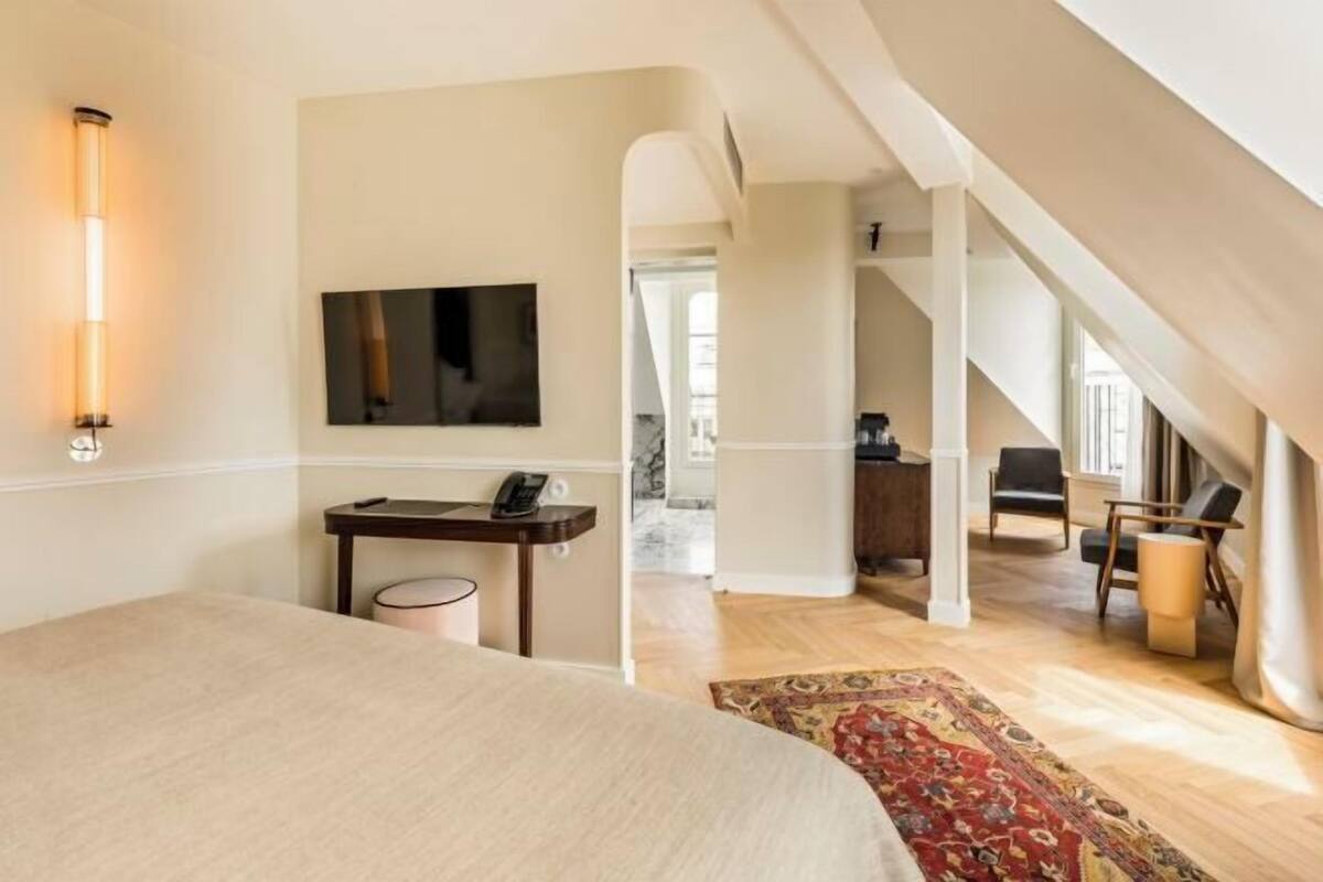 Un dormitorio con cama, mesita de noche, lámpara, televisor y vistas a un comedor con sillas y mesa.