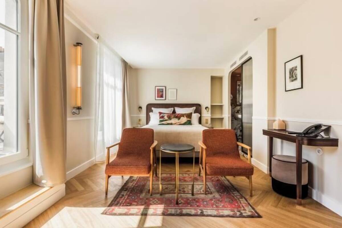 A hotel room with a bed, two armchairs, a small table, a bedside table, a lamp, a rug, and a window with curtains.