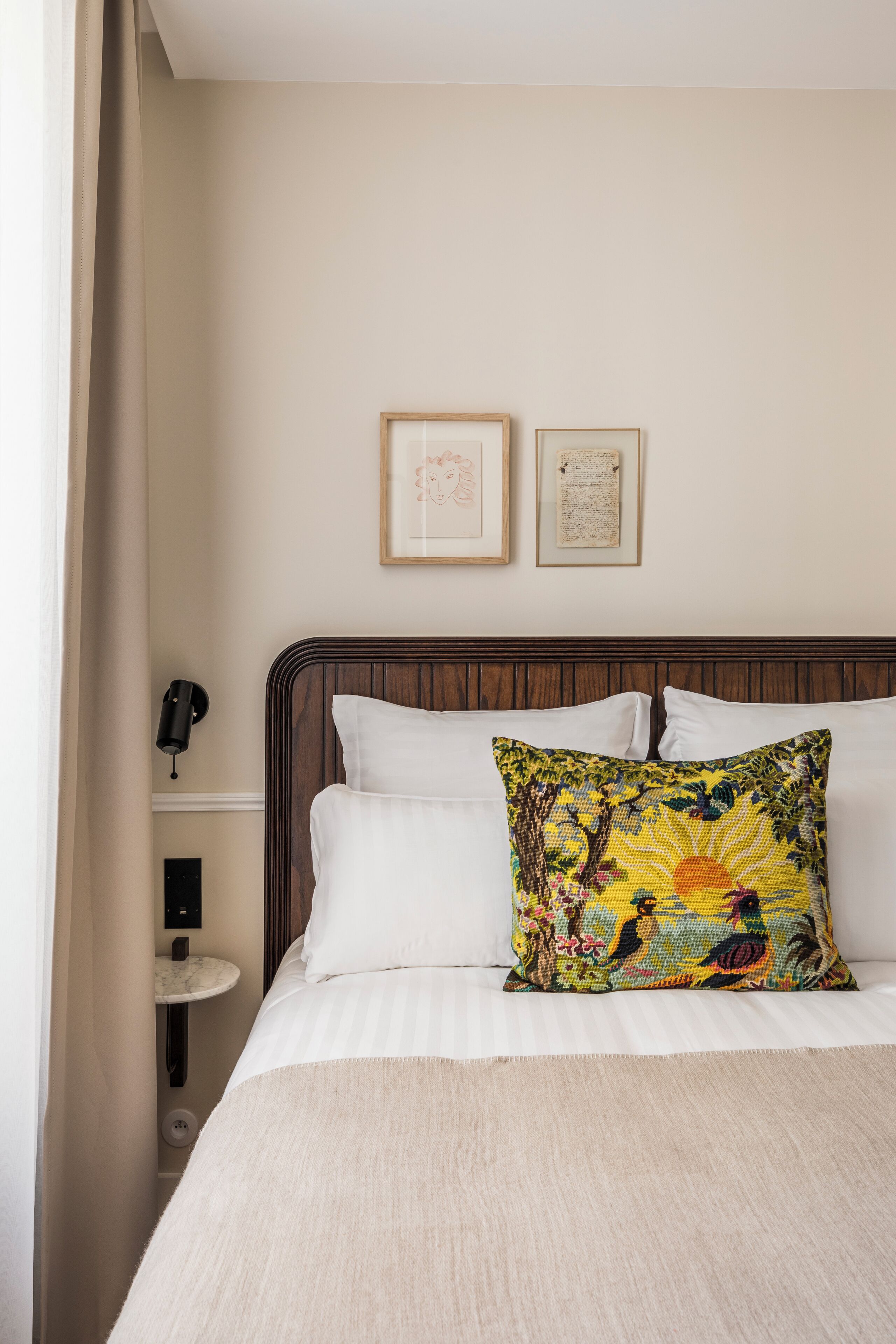 Una habitación de hotel con una cama, una mesita de noche, una lámpara de pared y cuadros enmarcados.