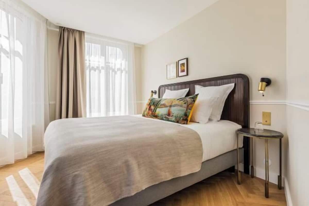 A hotel room with a large bed, a bedside table, a wall-mounted lamp, and a window with curtains.