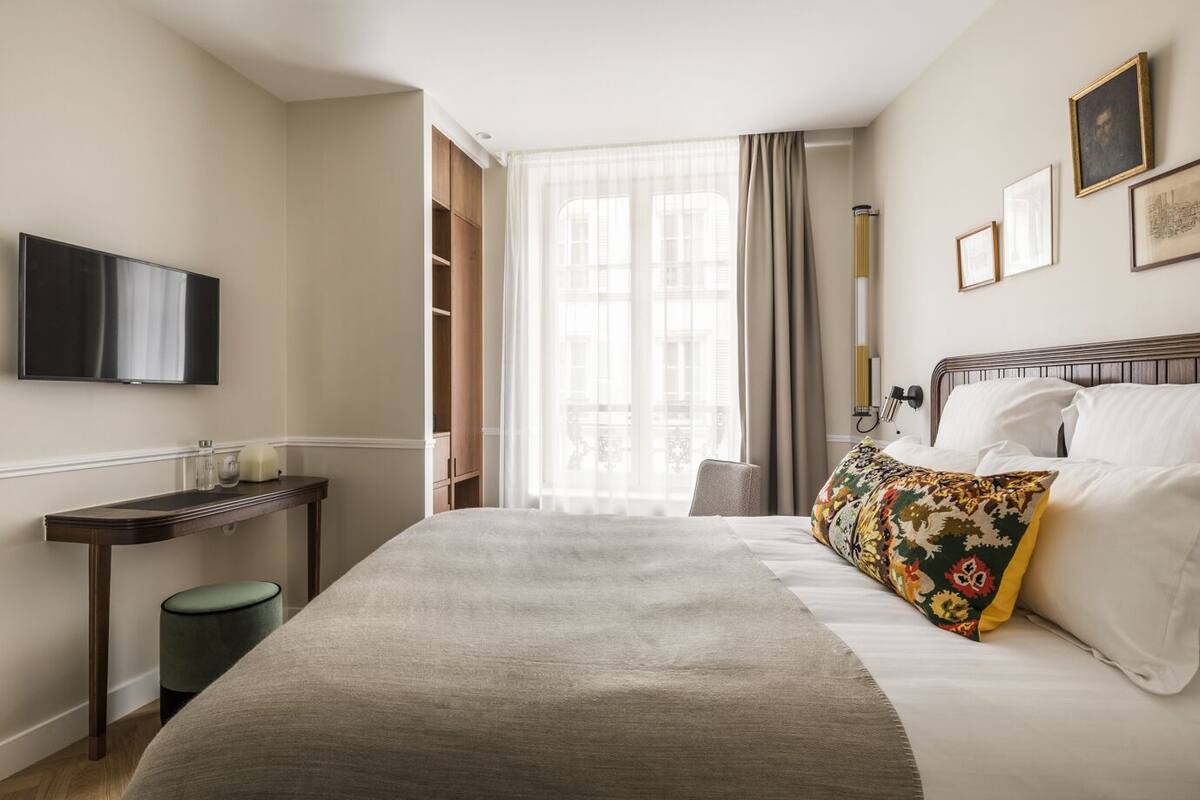 A hotel room with a bed, a TV mounted on the wall, a bedside table, and a window with curtains.