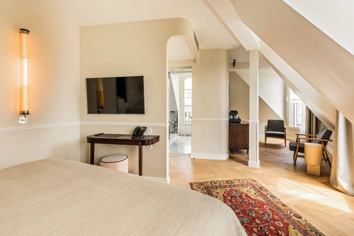 A bedroom with a bed, a desk with a phone, a TV, and a large window.