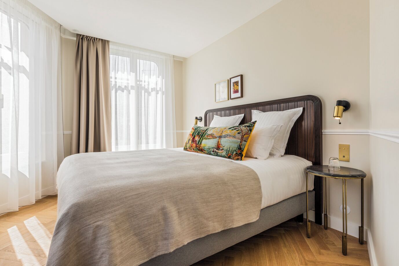 Una cama bien hecha con un edredón gris y una almohada colorida, una mesita de noche con una lámpara, y una ventana con cortinas translúcidas.