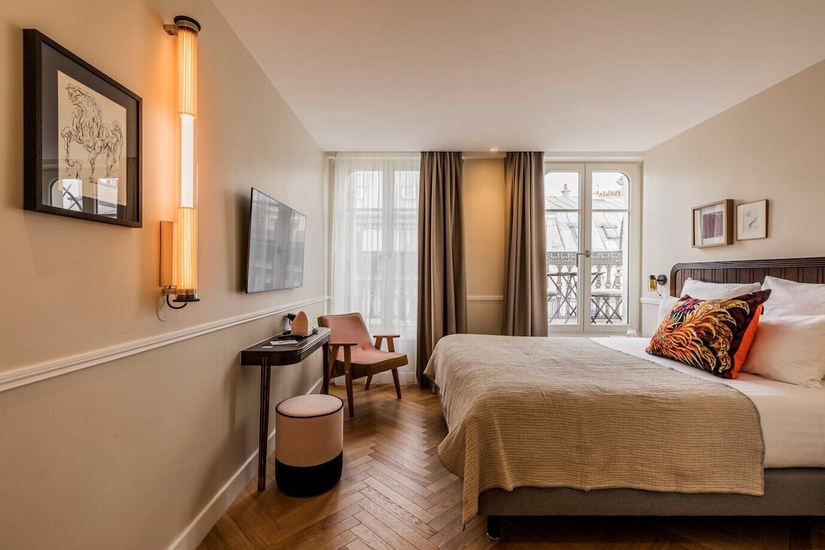 A hotel room with a bed, a desk, a chair, a waste bin, and a window with curtains.