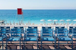 Un área de descanso frente a la playa con sillas azules y sombrillas a rayas, con vista al océano.