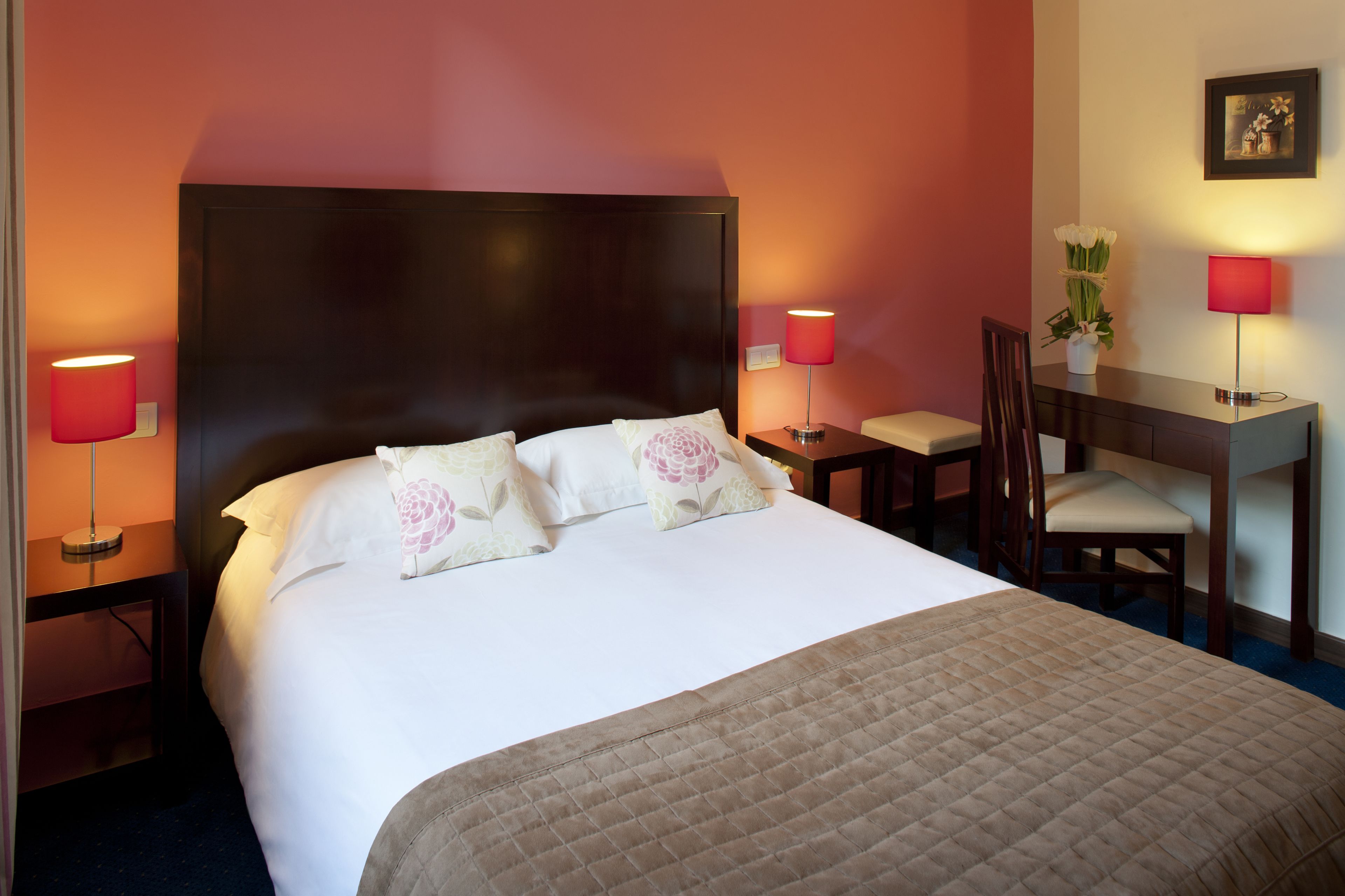 A hotel room with a large bed, two bedside tables with lamps, a desk with a chair, and a small potted plant.