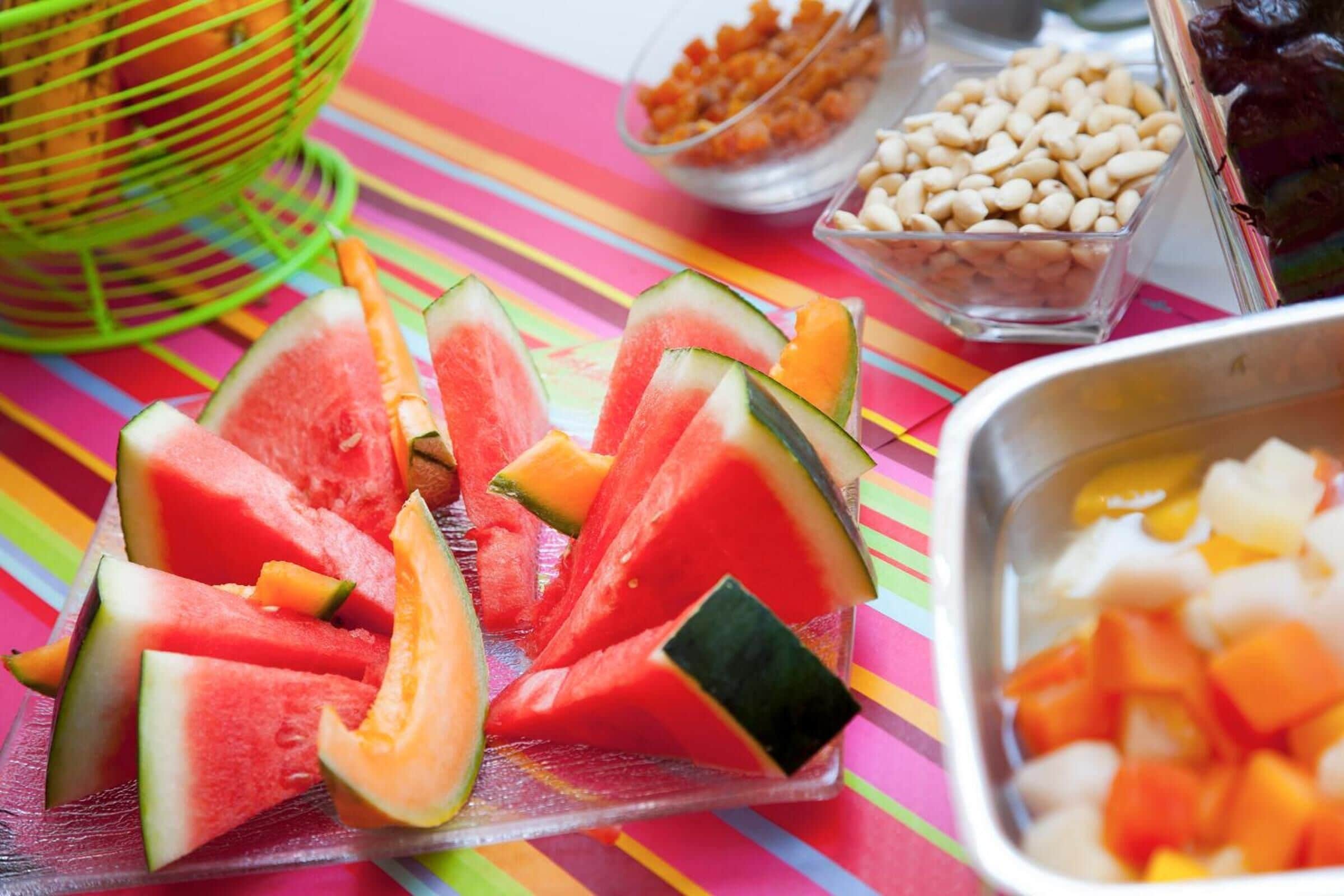 Een kleurrijke tafel met diverse soorten fruit en noten, waaronder watermeloen, meloen en pinda's.