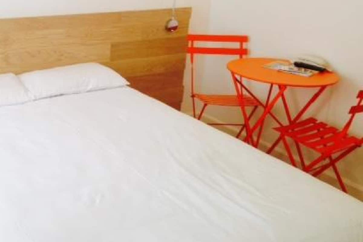 A bed with white bedding and a wooden headboard, a small orange table with a chair, and a red chair.