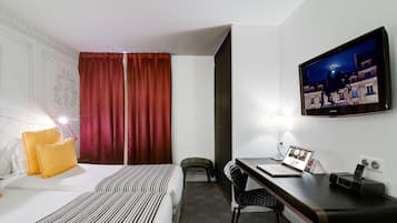 A hotel room with a bed, a desk with a laptop, a chair, a TV, and a window with curtains.
