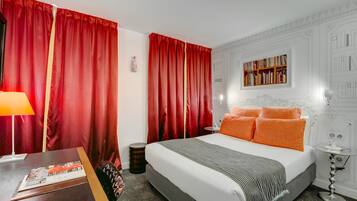 Habitación de hotel con una cama grande, cortinas rojas, una mesita de noche con lámpara, una pequeña mesita auxiliar y una estantería sobre la cama.
